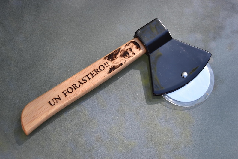 May include: A wooden pizza cutter shaped like an axe with a black handle and a silver blade. The handle is engraved with the text "UN FORASTERO!!"