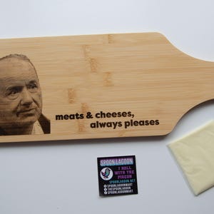 May include: A light-coloured bamboo serving board with a handle and a hole for hanging. It features a laser-engraved portrait of a man on the left and the text "meats & cheeses, always pleases" in black. A small square sticker and a slice of cheese are nearby.