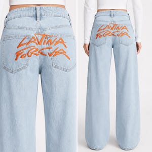 May include: Light blue denim jeans featuring a wide-leg silhouette and a high-rise waist. The back of the jeans displays the embroidered orange text "LAVINA FOREVA" across the two back pockets. The design includes standard belt loops and pockets.