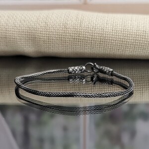 May include: A silver chain bracelet with a braided design. The bracelet is on a reflective surface.