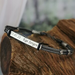 May include: A silver bracelet with a black braided cord and a silver plate engraved with the text "23.07.21 Ege Sergen".