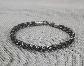 Könnte beinhalten: Ein geflochtenes Armband mit Silberverschluss. Das Armband besteht aus verschlungenen schwarzen und silbernen Strängen, wodurch ein strukturiertes, seilartiges Aussehen entsteht. Das Armband wird auf einer grauen Oberfläche präsentiert.