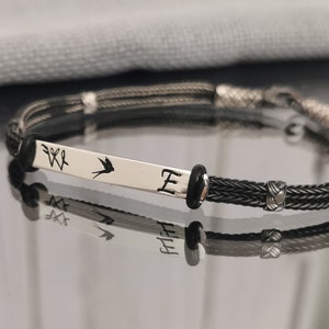 May include: A black braided bracelet with a silver charm that has a bird and the letter "E" engraved on it.
