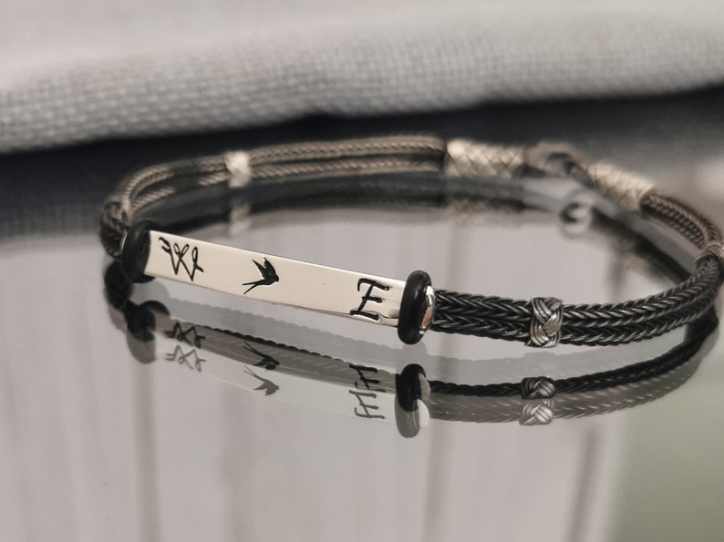 May include: A black braided bracelet with a silver charm that has a bird and the letter "E" engraved on it.