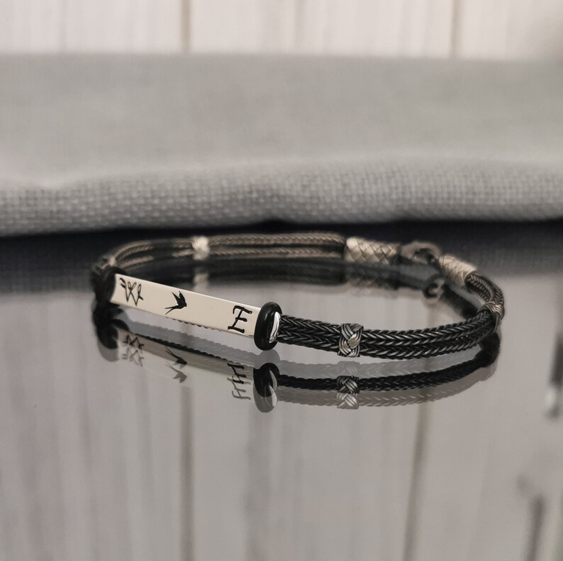 May include: A black braided bracelet with a silver charm featuring a bird and the letter "E".