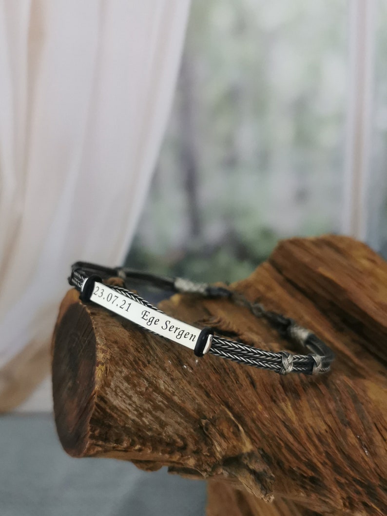May include: A silver bracelet with a black braided cord and a silver plate engraved with the text "23.07.21 Ege Sergen".