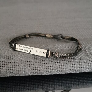 May include: A black braided bracelet with a silver charm that reads "Ik hou van je tot de maan en terug" and "BAE! S♡F".