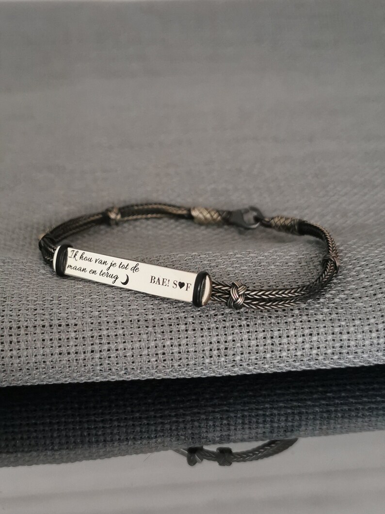 May include: A black braided bracelet with a silver charm that reads "Ik hou van je tot de maan en terug" and "BAE! S♡F".