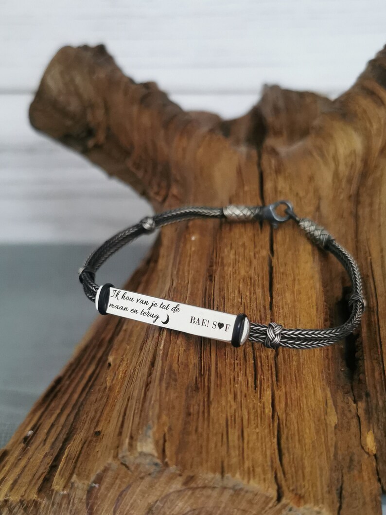May include: A silver bracelet with a black braided cord and a silver rectangular tag engraved with the text "Ik hou van je tot de maan en terug" and "BAE! S♡F".