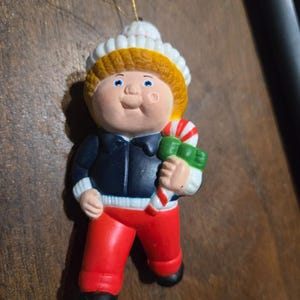 May include: A hanging ornament shaped like a child with blonde hair wearing a white knit hat, a dark blue jacket, and bright red pants. The figure holds a green gift box tied with a red and white striped candy cane ribbon, suspended by a gold string.
