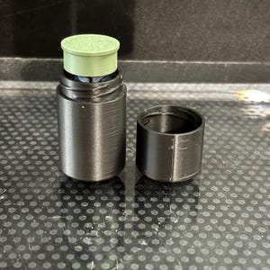Op de afbeelding: Een zwarte cilindrische container met een apart deksel. De container heeft een lichtgroene bovenkant. De items staan op een gestructureerd oppervlak. De container wordt waarschijnlijk gebruikt voor opslag of afgifte.