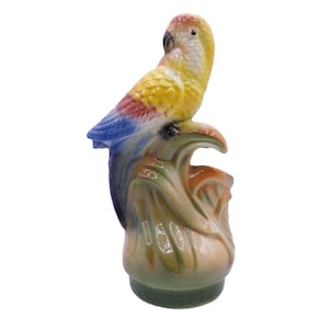 Puede incluir: Una figurita de loro de cerámica pintada en amarillo, azul, rojo y verde. El loro está posado en una rama verde con hojas y forma parte de un jarrón.