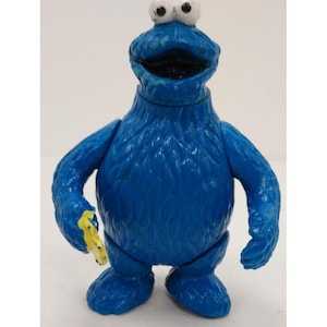 May include: A blue Cookie Monster figurine with white eyes and a wide open mouth. The figure is holding a yellow object in its right hand. The toy is a classic character from the children's television show Sesame Street.