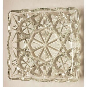 May include: A clear glass square dish with a cut glass design. The dish has a scalloped edge.