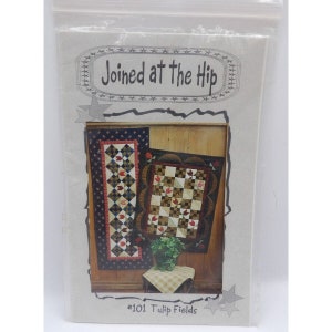 May include: A quilting pattern titled "Joined at the Hip" with the pattern number #101 Tulip Fields. The pattern features two quilt designs, one a long runner and the other a square wall hanging. Both quilts feature a red, white, and blue colour scheme with a floral design.