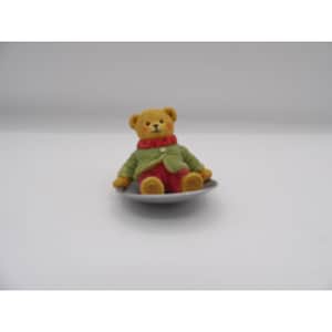 May include: A small, yellow teddy bear wearing a green sweater and red pants sits on a white saucer.