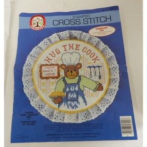 May include: A stamped cross stitch kit featuring a brown bear wearing a chef's hat and holding a pie. The design includes the text "Hug the Cook" and "Home Sweet Home". The kit is complete with everything needed to complete the project, including a 10 1/2 inch hoop.