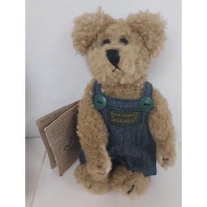 May include: A small, brown teddy bear wearing blue denim overalls with green buttons. The overalls have a brown patch with the text "Davy's Country Bearwear".