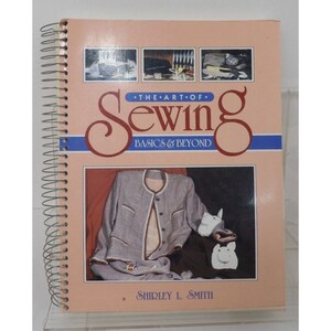 Op de afbeelding: Een roze spiraalgebonden boek getiteld "The Art of Sewing Basics & Beyond" door Shirley L. Smith. De omslag toont een foto van een grijze trui, een wit konijn en een wit varken.