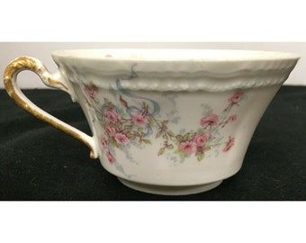 May include: A white porcelain teacup with a gold rim and handle. The cup is decorated with a floral pattern of pink roses and blue ribbons.
