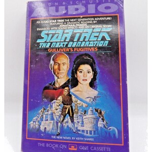 Puede incluir: Un casete de audio de Star Trek: La Nueva Generación de color morado y blanco con el libro "Los Fugitivos de Gulliver" de Keith Sharee. La portada presenta una foto de Patrick Stewart y Marina Sirtis.