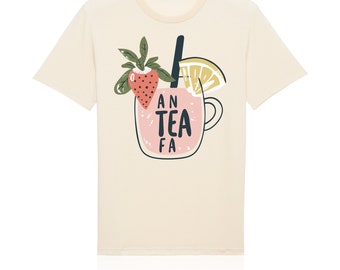 May include: A cream-colored short-sleeve t-shirt featuring a graphic of a pink drink in a glass mug. The design includes a strawberry, a lemon slice, and a black straw, with the text AN TEA FA printed in black letters on the front of the mug.