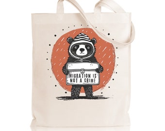 May include: A white canvas tote bag with a black and white illustration of a panda bear wearing a striped hat and holding a sign that says "Migration is not a crime". The panda is standing in front of a red circle with black dots.