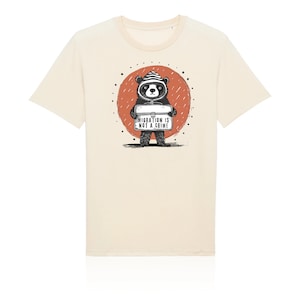 Peut inclure: Un t-shirt beige avec une illustration en noir et blanc d'un ours portant un chapeau et tenant une pancarte qui dit "La migration n'est pas un crime". L'ours se tient devant un cercle rouge avec des gouttes de pluie.
