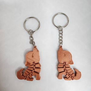 May include: Two copper-colored 3D printed dog keychains with silver key rings and chains. The dogs are in a sitting position with their heads tilted up.