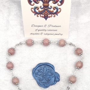 Peut inclure: Un chapelet composé de perles rose poudré avec des capuchons en métal argenté, un crucifix argenté et une médaille représentant une figure. Un sceau en cire bleue avec une fleur de lys est présent, et une carte indique Rosaries by Marcie.