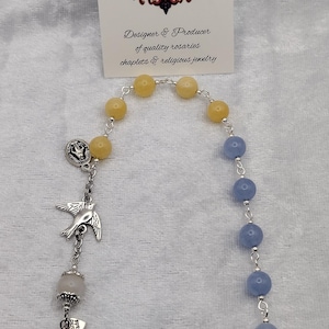 May include: A rosary with yellow and blue beads, silver accents, and a cross pendant. The rosary includes a dove charm and a St. Michael medal. The card reads "Rosaries by Marcie" and "Designer & Producer of quality rosaries chaplets & religious jewelry."