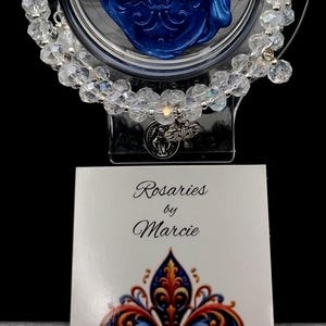 Op de afbeelding: Transparante kristallen rozenkransarmband met zilverkleurige accenten, gepresenteerd in een transparante houder met een blauw fleur-de-lis-ontwerp. Een kaart leest "Rosaries by Marcie" met een fleur-de-lis-ontwerp.