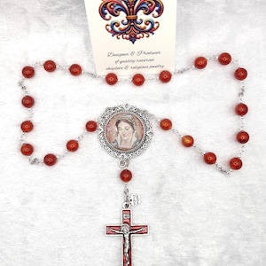 May include: A rosary with red beads and a silver chain. The rosary features a central medallion with a religious image and a red crucifix. The card behind the rosary reads "Rosaries by Marcie."