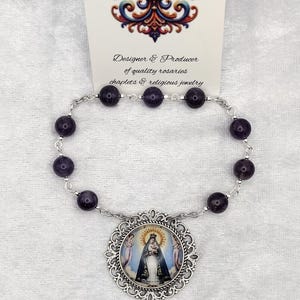 May include: A rosary with dark purple beads and a silver-toned crucifix. The centerpiece features an image of a religious figure. The image includes a card with the text "Rosaries by Marcie" and a fleur-de-lis design.