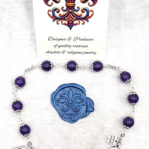 Peut inclure: Un chapelet de Marcie avec des perles violettes, une croix bleue et un sceau bleu fleur-de-lis. Le chapelet comprend un crucifix argenté et une médaille de Saint Christophe. Le texte sur la carte indique "Rosaries by Marcie".