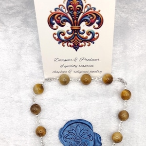 Peut inclure: Un chapelet avec des perles marron et une chaîne argentée, comprenant un crucifix et une médaille de Saint Christophe. Un sceau de cire bleu avec un motif de fleur de lys est également présent. La carte indique "Rosaries by Marcie".