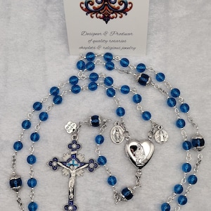 Op de afbeelding: Een rozenkrans met blauwe kralen en zilverkleurige metalen accenten. De rozenkrans heeft een kruisbeeld, een hartvormige hanger en een fleur-de-lis-ontwerp. De afbeelding bevat ook een kaart met de tekst "Rosaries by Marcie."