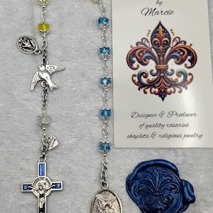 May include: A handmade rosary with yellow and blue beads, a silver cross with a blue border, and a dove charm. The image includes a card that reads "Rosaries by Marcie" and a blue wax seal.