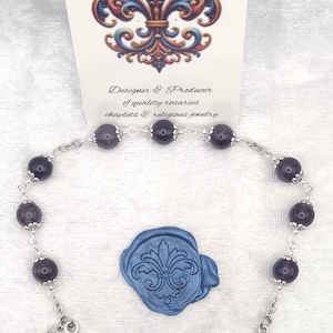 Peut inclure: Un chapelet composé de perles violet foncé, de maillons en métal argenté, d'un crucifix et d'une médaille représentant une figure avec une mitre. La carte indique Rosaries by Marcie, Designer & Producer of quality rosaries chaplets & religious jewelry.