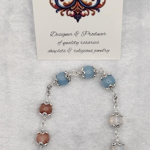 May include: A handmade rosary with light blue and peach-colored beads, silver accents, and two religious medals. The rosary is displayed with a card that reads "Rosaries by Marcie" and "Designer & Producer of quality rosaries, chaplets & religious jewelry."