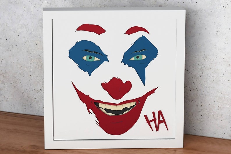 May include: A white background with a red, blue, and black illustration of a Joker's face. The Joker's eyes are blue and the mouth is open in a wide smile. The text "HA" is written in red below the mouth.