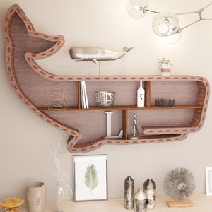 Puede incluir: Una estantería de pared con forma de ballena de madera, con múltiples compartimentos. La estantería es marrón con un borde cosido y contiene objetos decorativos como libros, una figura de ballena de metal y un cuenco decorativo.