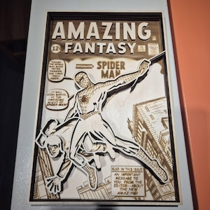 May include: A framed wooden artwork featuring a comic book cover of Spider-Man. The title "AMAZING FANTASY" is at the top, with Spider-Man in action. The artwork is in shades of brown and white, with text bubbles and building details.
