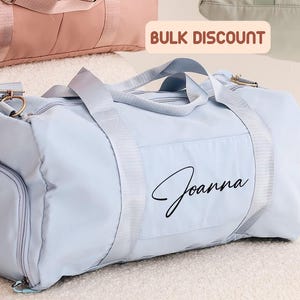 May include: Three duffel bags in pastel colors: pink, mint green, and light blue. The light blue bag has the name "Joanna" printed on it. The bags have shoulder straps and side pockets. The text "BULK DISCOUNT" is visible.