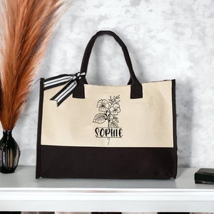 May include: A beige and black canvas tote bag with black and white striped ribbon handles. The bag features a floral design and the name "Sophie" printed on the front.