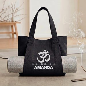 May include: A black canvas tote bag featuring a white Om symbol and the name AMANDA printed in white text. The bag has a horizontal strap across the front that holds a rolled-up light gray textured yoga mat securely in place.