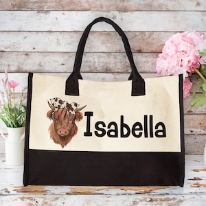 K&ouml;nnte beinhalten: Eine beige und schwarze Tragetasche mit einem braunen und wei&szlig;en Kuhkopfdesign und dem Namen "Isabella" in Schwarz gedruckt.