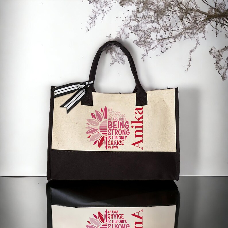 May include: A beige and black tote bag featuring a sunflower design and the text "We don't know how strong we are until being strong is the only choice we have". The bag is personalised with the name "Anika".