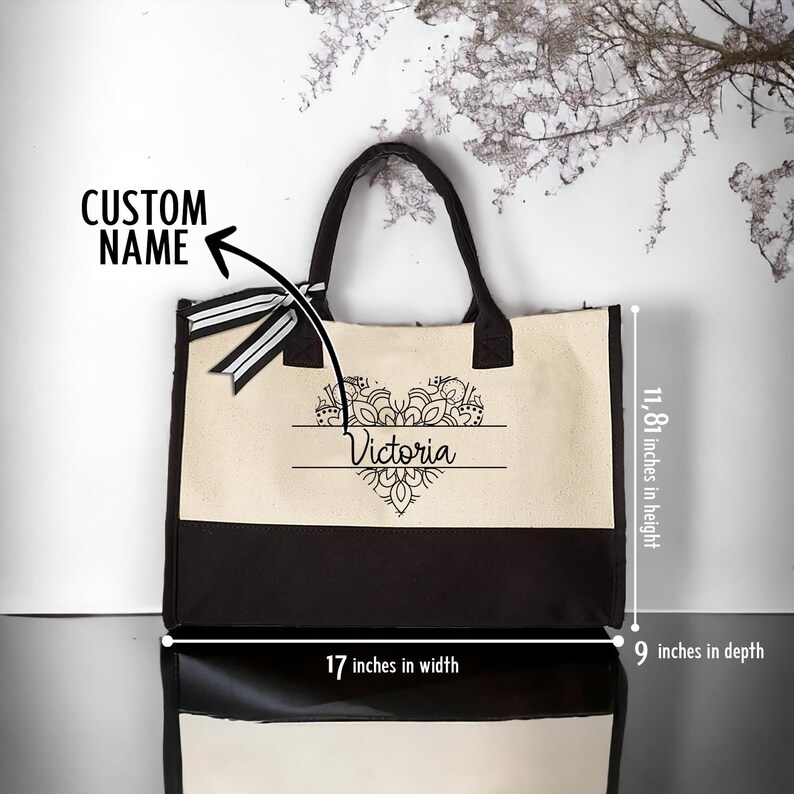 May include: A black and white canvas tote bag with a black handle. The bag features a white heart design with a mandala pattern and the name "Victoria" written in black. The bag measures 17 inches wide, 11.81 inches high, and 9 inches deep.