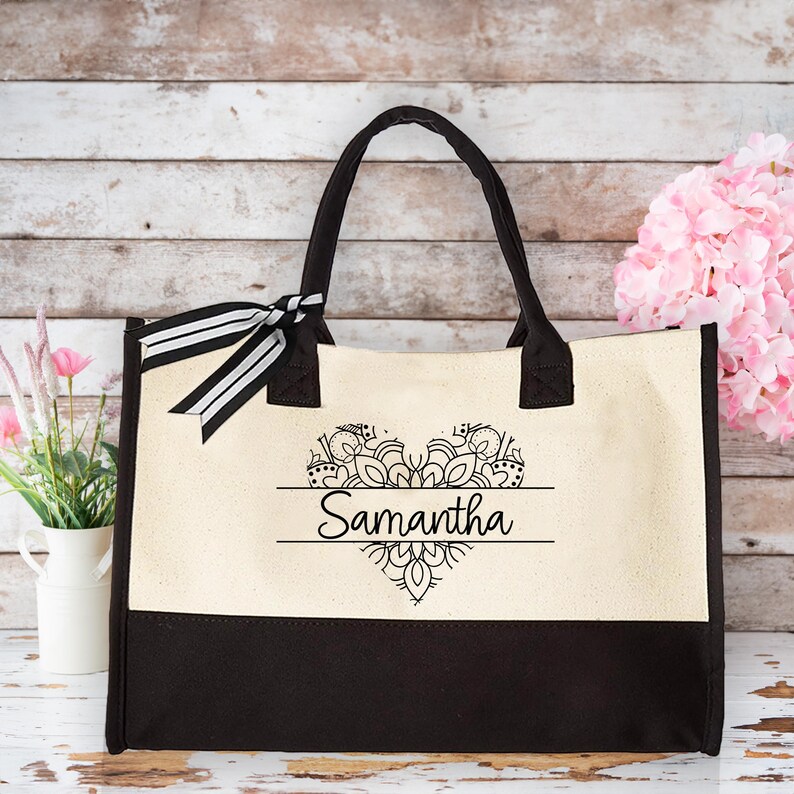 May include: A black and white tote bag with a black and white mandala heart design and the name "Samantha" printed on it. The bag has black handles and a black bottom.